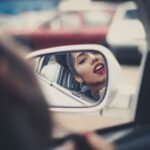 Dark haired woman applying lipstick in side view car mirror.