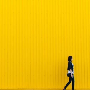 Dark haired woman walking along a yellow wall.